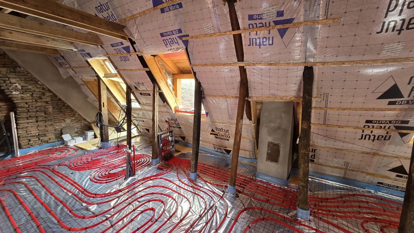 Mansardă cu serpentine roșii de încălzire în pardoseală pe folie reflectorizantă, printre stâlpii acoperișului